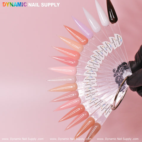 A fan display of Dynamic Nail Supply's (BIAB) Rubber Base Gel (Builder Gel in a Bottle) Number 13 to 29 collection showcases a gradient of colors from pale pinks to deep brown, featuring both glossy and muted shades. Each sample is labeled and attached to a metal ring, set against a soft pink background with the display held by a gloved hand.