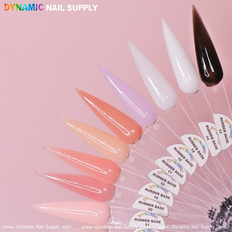 A fan of pointed nail polish samples from Dynamic Nail Supply graces a pink background. Showcasing the (BIAB) Rubber Base gel, the samples include shades from dark brown to delicate hues of pink, peach, light purple, white, and beige. Each is labeled with numbers 13 to 29 from the Builder Gel in a Bottle collection.