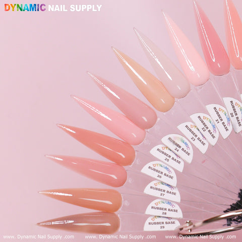 A fan-shaped display features pointed gel polish samples in a gradient of nude to pink shades from the (BIAB) Rubber Base Gel (Builder Gel in a Bottle) Number 13 to 29 collection. Each swatch is labeled with color names and numbers. The soft pink background accentuates Dynamic Nail Supply's logo at the top and bottom, showcasing their signature nails gel polish collection.