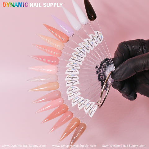 A display features an array of shades from the (BIAB) Rubber Base Gel (Builder Gel in a Bottle) Number 13 to 29 collection by Dynamic Nail Supply, beautifully arranged on long, pointed swatches. The spectrum ranges from light pinks and nudes to deep browns and blacks. A gloved hand elegantly holds the fan-shaped arrangement against a pink backdrop with the brand name, Dynamic Nail Supply, prominently displayed at the top.