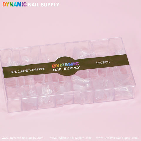 A transparent plastic box containing 550 clear 90's Curve Down Tips in square shapes is set against a light pink background. The label on the box showcases vibrant text that reads "Dynamic Nail Supply." Below, www.DynamicNailSupply.com appears on the pink surface, emphasizing their easy application.