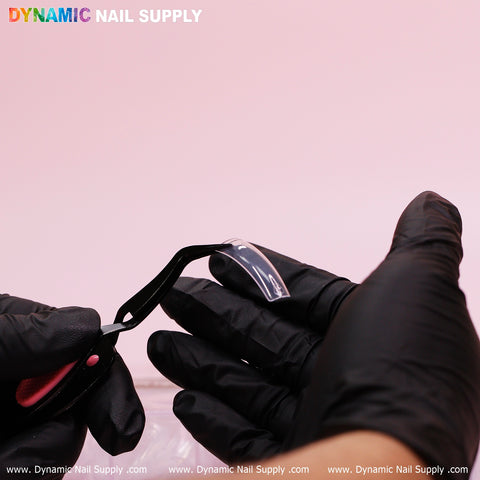 A person wearing black gloves holds a 90's Curve Down Tip from Dynamic Nail Supply using curved tweezers against a pink background. The brand name DYNAMIC NAIL SUPPLY is displayed at the top, with the website www.DynamicNailSupply.com repeated at the bottom, highlighting their collection of easy-to-apply, clear square tips available in 10 sizes with 550 tips per set.