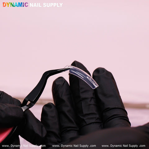 The image showcases a gloved hand using tweezers to expertly hold a 90'S Curve Down Square translucent nail tip. The soft pink background highlights the colorful letters spelling out "Dynamic Nail Supply" at the top, with the website repeated along the bottom edge. These clear tips, which come in 10 different sizes with a total of 550 tips, are simple to apply.