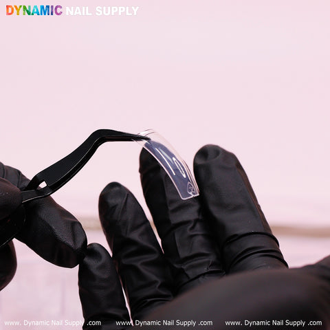 A person wearing black gloves is holding an easy-to-apply clear 90's Curve Down Square nail tip with black tweezers against a pinkish background. The image highlights the quality of Dynamic Nail Supply, prominently displaying the brand name and website at both the top and bottom.