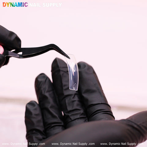 A person wearing black gloves uses tweezers to hold a curved, transparent nail tip from the 90'S Curve Down Tips collection. These tips, which are easy to apply, stand out prominently against a gently blurred background. The brand Dynamic Nail Supply is featured at the top of the image, while light reflects off the clear square tip positioned at the center.