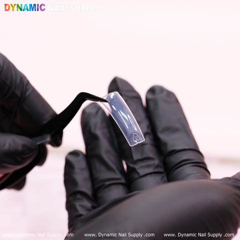 A gloved hand delicately holds the easy-to-apply 90's Curve Down Tips - Square with black tweezers, while another gloved hand provides support beneath. On a soft pink background, "Dynamic Nail Supply" is elegantly displayed at the top, with a website URL in pristine white at the bottom.