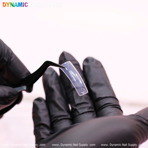 A close-up image shows gloved hands using a black tweezer to hold a clear, glossy nail tip, designed for easy application in any style. The background is blurred, with the top left corner featuring the Dynamic Nail Supply logo in colorful text. For more information on the 90's Curve Down Tips - Square (10 sizes, 550 tips), visit www.DynamicNailSupply.com.