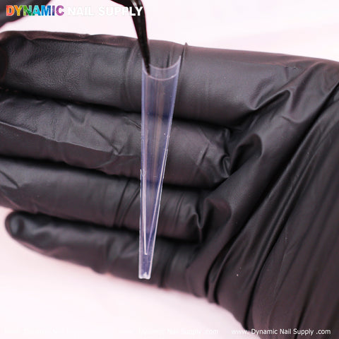 A black-gloved hand elegantly grasps a clear 4XL Super Long Stiletto nail tip with tweezers. The vertically positioned tip showcases its sleek curvature and impressive length, set against a blurred background. Vibrant text at the top left corner highlights DYNAMIC NAIL SUPPLY, emphasizing their elegance.