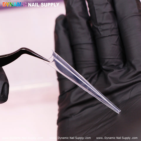 Against a soft pink backdrop, a gloved hand gently holds one of Dynamic Nail Supply's 4XL Super Long Stiletto Nail Tips using black tweezers. Text is displayed at the bottom, while the vibrant logo of Dynamic Nail Supply is prominently featured at the top.