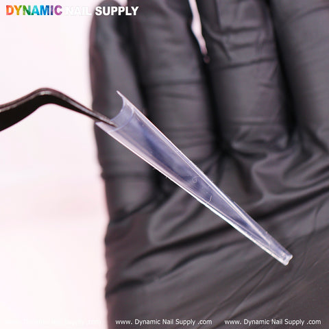 A gloved hand uses tweezers to hold a translucent, elongated 4XL Super Long Stiletto Nail Tip from Dynamic Nail Supply, which gradually tapers to a point. The brand name is displayed at the top of the image, with the website name repeated at the bottom.