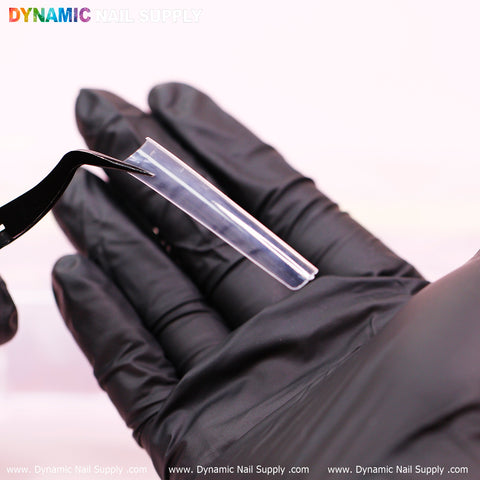 A person wearing black gloves holds a clear, durable 3XL Tapered Square Tip with tweezers. The blurred background features soft pink hues, with the brand name Dynamic Nail Supply displayed at the top. A website link, www.DynamicNailSupply.com, is visible at the bottom of the image.