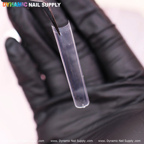 A gloved hand holds a clear, curved nail extension with tweezers against a blurred background. The black glove highlights the transparent 3XL Tapered Square Tips. The brand name, Dynamic Nail Supply, is displayed in colorful text at the top of the image.