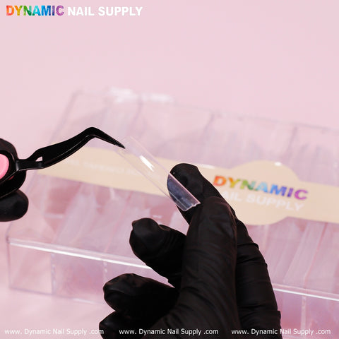 A person is carefully holding a durable and robust 3XL Tapered Square Tip from Dynamic Nail Supply with black-gloved hands, using tweezers. In the background, a plastic container labeled Dynamic Nail Supply is partially visible against a pink surface, seemingly filled with more clear tips from their latest 2024 release.