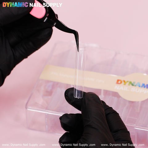 A person wearing black gloves uses tweezers to hold a 3XL Tapered Square Tip. In the background, there's a plastic container branded with Dynamic Nail Supply. The clear tip stands out against the soft pink background, promising durability and strength.