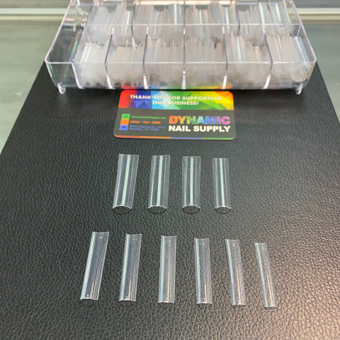 On a black textured surface, clear acrylic nail tips are arranged in two rows, highlighting their role in an enticing Acrylic Nail Starter Kit for professionals or beginners. In the background, an open container displays additional tips. Nearby, a colorful business card for Dynamic Nail Supply is placed, featuring contact details that any nail salon would find valuable.