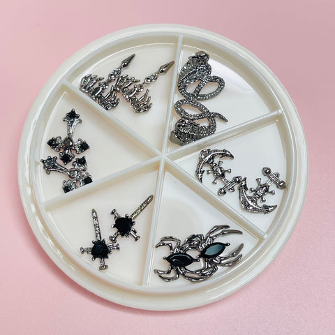 A round white container from Dynamic Nail Supply sits on a pink surface, containing an assortment of silver and black charms for nail art. Among the intricate designs are crowns, fleur-de-lis, a sword, and gothic lettering, each elaborately adorned with small black or clear stones. These items are part of the "12 pcs Unique Vintage Gothic Halloween Charms," featuring crosses, snakes, and spiders.