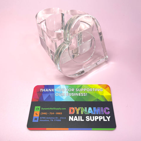 A CLEAR HEART SHAPE DAPPEN DISH sits on a pink surface, with a vibrant business card for Dynamic Nail Supply displaying a rainbow background in front of it. The card includes contact information and thanks customers for supporting their Liquid Monomer products.