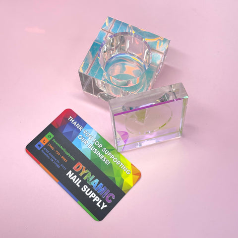 A business card for Dynamic Nail Supply is placed on a pink surface next to two transparent SQUARE SHAPE DAPPEN DISH containers. The card displays a colorful design with social media icons and a thank you message. The glass captures light, creating rainbow hues, ideal for presenting liquid monomer essentials.