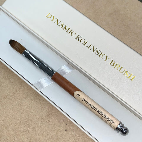 A close-up image features a brush from the Dynamic Nail Supply Acrylic Nail Starter Kit, ideal for professionals or beginners. The brush showcases a brown wooden handle and silver metal ferrule with fine bristles, all elegantly placed in a white box with "DYNAMIC KOLINSKY BRUSH" etched in gold. The simple brown backdrop is perfect for any nail salon setting.