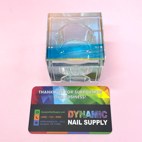 A SQUARE SHAPE DAPPEN DISH, evocative of crystal glass, contains a blue liquid monomer set against a pink background. In front is a vibrant business card from Dynamic Nail Supply, complete with contact information and a heartfelt message: Thank you for supporting our business!