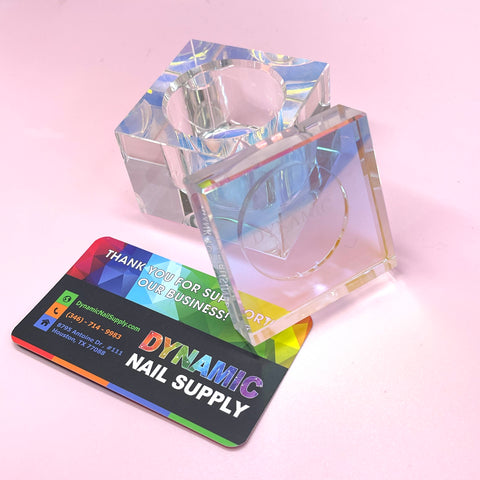 A stylish SQUARE SHAPE DAPPEN DISH featuring a cylindrical center is elegantly placed on a pink surface. Next to it sits a colorful business card for Dynamic Nail Supply, showcasing contact information along with a sincere thank you message for supporting our business.