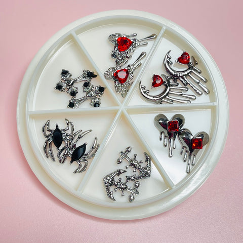 Introducing the Dynamic Nail Supply's 12 pcs Vintage Gothic Halloween Charms in red and black, ideal for nail art designs. These charms are presented on a round white dish featuring six compartments. Each compartment displays Gothic rhinestone designs, including jeweled spiders, hearts, and abstract shapes. Perfect for nail art designers seeking an edgy touch against a pale pink background.