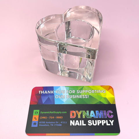 A dappen dish made of clear glass in a heart shape, from Dynamic Nail Supply, sits on a pink surface. In front, a vibrant business card highlights contact details such as phone number, website, and the Houston, TX address for Dynamic Nail Supply. The card reads: Thank you for supporting our business!