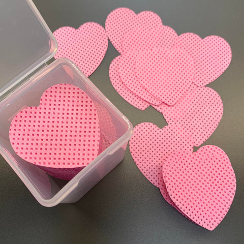 A transparent plastic container contains multiple pink, heart-shaped coasters with delicate grid patterns, resembling professional salon essentials from Dynamic Nail Supply. Additional coasters are artfully arranged outside the package on a dark surface, creating an appealing display similar to the 200 pcs Heart Shape Lint-free Wipes for Nails or Lashes.