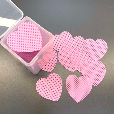 An open small transparent box from Dynamic Nail Supply, containing their 200 pcs Heart Shape Lint-free Wipes for Nails or Lashes, rests on a gray surface. Scattered around the box are several heart-shaped lint-free wipes that resemble non-woven fabric, adding a whimsical touch to what could be professional salon supplies. Each wipe features a pink heart design with black polka dots.