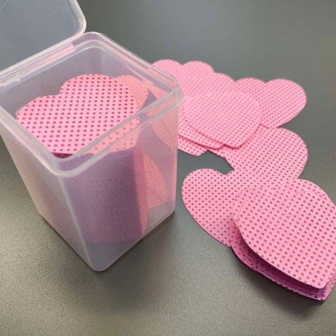 A clear plastic container holds several pink, heart-shaped cards with a dotted pattern, while more cards are scattered on a dark gray surface beside it. The 200 pcs Heart Shape Lint-free Wipes for Nails or Lashes by Dynamic Nail Supply are stacked neatly, highlighting their consistent heart shape and bold pink color.