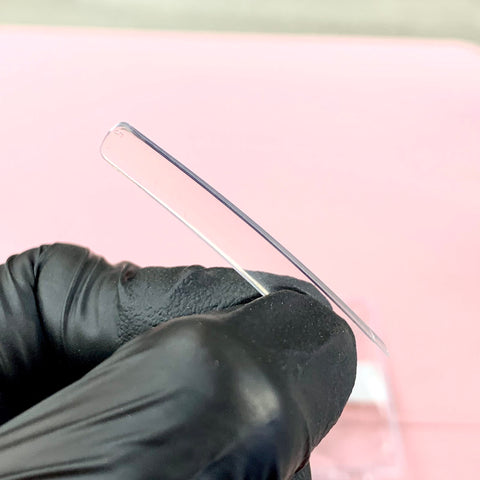 A gloved hand holds a thin, transparent 3XL Tapered Square Tip from Dynamic Nail Supply, viewed from the side. The durable and stronger tip is slightly curved and appears clear against a blurred pink background. The black glove provides a stark contrast to the delicate, glass-like material of the nail tip.