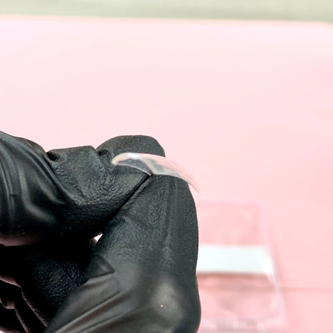 A gloved hand holds a thin, transparent, curved object that resembles the 3XL Tapered Square Tips from Dynamic Nail Supply. The softly blurred pink background highlights an indistinct transparent box nearby. The black glove offers stark contrast, enhancing the durable and stronger translucency of these new 2024 clear tips.