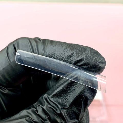 A gloved hand is holding a clear, thin plastic rectangle with slightly rounded edges against a pink background. The black textured glove suggests durability and strength, as if handling Dynamic Nail Supply's new 2024 3XL Tapered Square Tips. The object appears flexible and transparent, similar to the design of these clear tips.