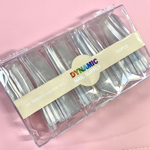 A clear plastic box holds 550 pieces of the durable and stronger 3XL tapered square nail tips. The packaging displays the brand name "Dynamic Nail Supply" in colorful text against a soft pink background.