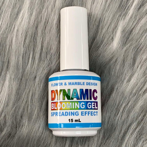 A bottle of Dynamic Nail Supply's Dynamic Blooming Gel, ideal for nail artistry, featuring a white cap is positioned on a gray, furry surface. The label prominently displays "Flower & Marble Design" at the top and "Spreading Effect" at the bottom. The name "Dynamic Blooming Gel" stands in bold letters, promising an exquisite marble effect with its 15 mL content.