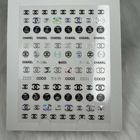 A sheet of chic silver holographic nail art stickers from Dynamic Nail Supply features the iconic designs of interlocking C's and the word "CHANEL." These Logo Stickers for nails art design - Md005 - easily peel off and adhere to your nails, adding a glamorous touch. The sheet is beautifully displayed against a plush white fur background.