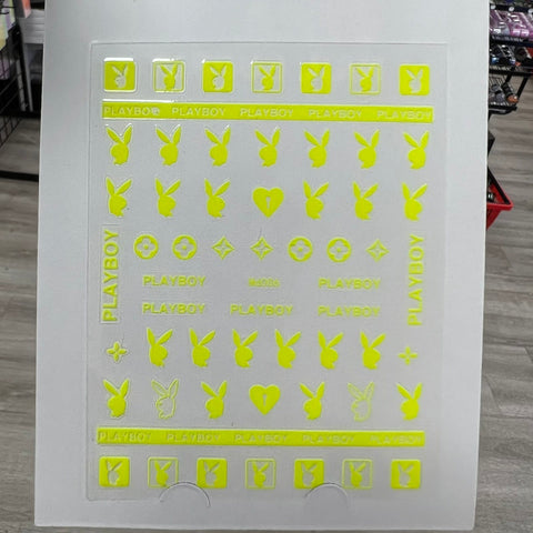 A sheet of Dynamic Nail Supply's Logo Sticker for Nail Art Design - Md006 - Yellow, featuring repeated Playboy bunny logos, the word PLAYBOY, hearts, and various abstract symbols on a transparent backing. Ideal for nail art enthusiasts, this vibrant collection is displayed on a wooden floor backdrop.