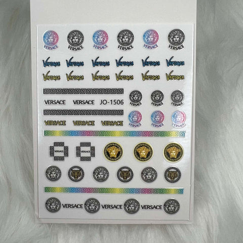A set of Logo Stickers for nail art design - JO-1506 in a Versace theme displayed on a white, furry background. These versatile peel-and-stick designs from Dynamic Nail Supply feature colorful and monochrome Medusa logos, various styles of Versace text, intricate Greek key patterns, and rows of circle motifs in black, gold, and multicolor hues.