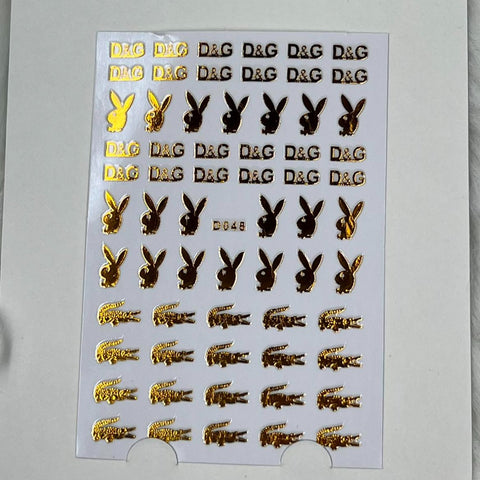 The Logo Sticker for nails art design - D048 - Gold by Dynamic Nail Supply showcases rows of stunning gold metallic stickers featuring patterns of D&G logos, bunny silhouettes, and small crocodile designs. With a total of 55 peel-and-stick designs that shine brilliantly on a white background, these are perfect for nail art enthusiasts.