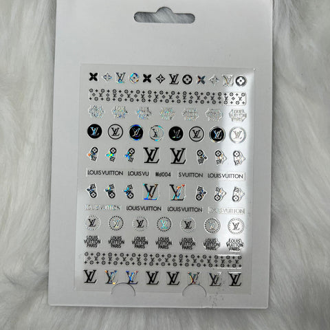 A sheet titled "Logo Sticker for nails art design - md004 - Silver Holographic" by Dynamic Nail Supply, featuring an assortment of logos, symbols, and the brand name Louis Vuitton in various fonts and styles. These silver peel-off nail art stickers are meticulously arranged in rows against a white fluffy backdrop.
