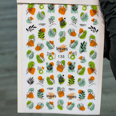 A sheet featuring the FALL Nail Designs Stickers for Fall Season 2021 by Dynamic Nail Supply showcases vibrant leaf and plant stickers in various styles and sizes, with rich shades of orange, green, and black. With the label "leaves" and "135," it is ideal for fall nail designs. The display on a white background is perfectly captured as it's held upright by a person's hand.