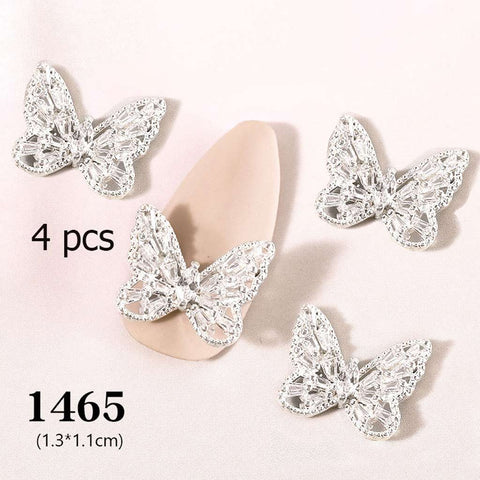 On a beige surface, four detailed 3D silver butterfly charms from Dynamic Nail Supply are showcased. One of these charming ornaments is used to decorate a nail art design sample tip. The description highlights "4 pcs 3D Butterfly Charm for Nails Art Design (code 1465)," specifying the quantity and dimensions of each charm at 1.3 cm by 1.1 cm, available through Dynamic Nail Supply.