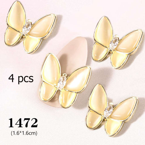 A set of four 3D butterfly charms, featuring gold designs with cream-colored enamel wings and a central rhinestone, is displayed on a light surface. These butterfly-shaped pieces measure 1.6*1.6 cm each and are perfect for enhancing your nail art design. Available from Dynamic Nail Supply under product code 1472, this set adds an elegant touch to your nails.