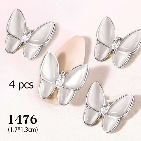 On a light background, four white butterfly charms with silver edges from Dynamic Nail Supply can be seen. The text indicates "4 pcs 3D Butterfly Charm for Nails Art Design (code 1476)" in black font, catering to nails art enthusiasts. A subtle pink shape appears beneath the butterflies.