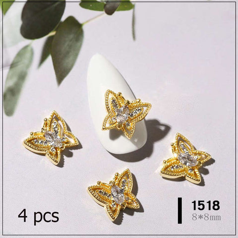 Displayed on a pale, glossy surface, this set of four ornate butterfly nail charms from Dynamic Nail Supply features a gold-colored floral design with clear rhinestones at the center. A green leaf is partially visible in the top left corner. Enhance your nail art design with these stunning pieces. Text: 4 pcs and 1518 8*8mm at the bottom.