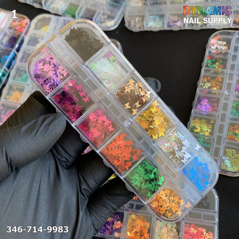 A gloved hand holds a rectangular box filled with vibrant nail art decorations, ideal for any nail design enthusiast. Glitter stars and shapes in pink, orange, green, and blue embellish each compartment. The box is labeled with the product name "NAIL DESIGN GLITTER 1" from Dynamic Nail Supply and features the phone number 346-714-9983 prominently.