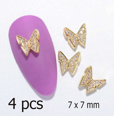 Four stunning 3D butterfly charms, crafted in gold and encrusted with crystals, are elegantly displayed. One charm embellishes a purple nail swatch, while the remaining three lie nearby, ready to elevate any nail art design. The accompanying text highlights "4 pcs" and specifies dimensions of 7 x 7 mm. Offered by Dynamic Nail Supply for creative expressions.
