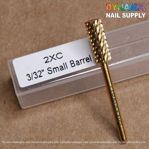 A gold 2XC Double Extra Coarse Drill Bit - Small Barrel Carbide, ideal for removing gel or acrylic nails, branded as Dynamic Nail Supply, lies on a beige surface. Behind it sits a clear plastic case with a white label stating "2XC 3/32” Small Barrel." The vibrant Dynamic Nail Supply logo decorates the top right corner.