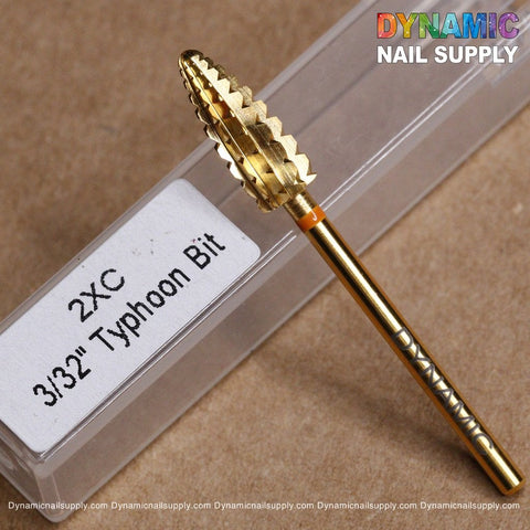 Close-up of a gold-colored 2XC Typhoon Bit, a double extra coarse carbide drill bit ideal for removing acrylic or gel polish, resting on a brown surface. It is accompanied by a clear plastic case with a white label. The top right corner features the Dynamic Nail Supply logo in vibrant multicolored letters.
