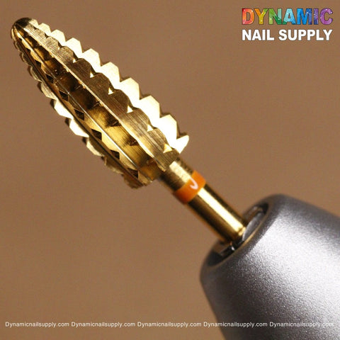 A close-up of the 2XC Typhoon Bit by Dynamic Nail Supply, a gold-colored, textured drill bit featuring a conical shape with serrated edges, designed for removing acrylic or gel polish. It is securely housed in a metallic holder. Located in the upper right corner is the vibrant Dynamic Nail Supply logo. The warm tone of the soft brown background enhances the overall presentation.